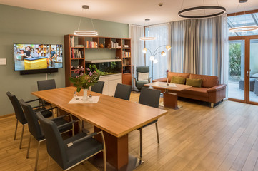 Blick in den wohnlich eingerichteten hellen Aufenthaltsraum mit Holzfußboden, indirekter Beleuchtung, einem großen Tisch mit Stühlen, Sofa, Regal und großem Monitor im Diakonie Hospiz Volksdorf in Hamburg-Volksdorf. Blick in den wohnlich eingerichteten hellen Aufenthaltsraum mit Holzfußboden, indirekter Beleuchtung, einem großen Tisch mit Stühlen, Sofa, Regal und großem Monitor im Diakonie Hospiz Volksdorf in Hamburg-Volksdorf.