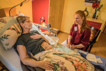 Eine Fachpflegerin in weinroter Funktionskleidung sitzt neben dem Pflegebett im Gästezimmer einer Hospizgästin und unterhält sich mit ihr im Diakonie Hospiz Volksdorf in Hamburg-Volksdorf. Eine Fachpflegerin in weinroter Funktionskleidung sitzt neben dem Pflegebett im Gästezimmer einer Hospizgästin und unterhält sich mit ihr im Diakonie Hospiz Volksdorf in Hamburg-Volksdorf.